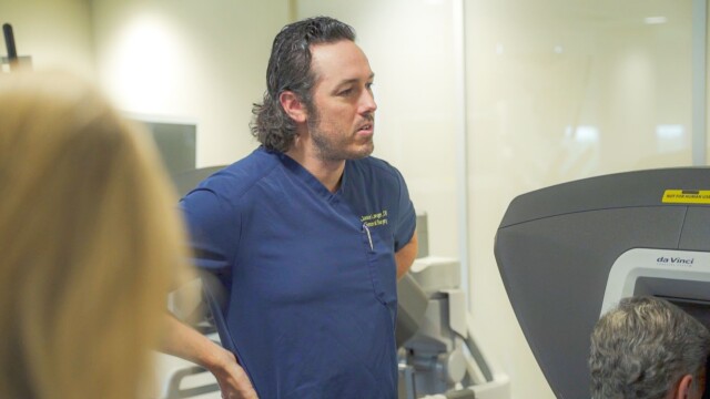 A man in blue medical scrubs, possibly a Phoebe Surgeon, stands with hands on hips, watching a person seated at a da Vinci medical machine in a clinical setting. Another figure is blurred in the foreground.